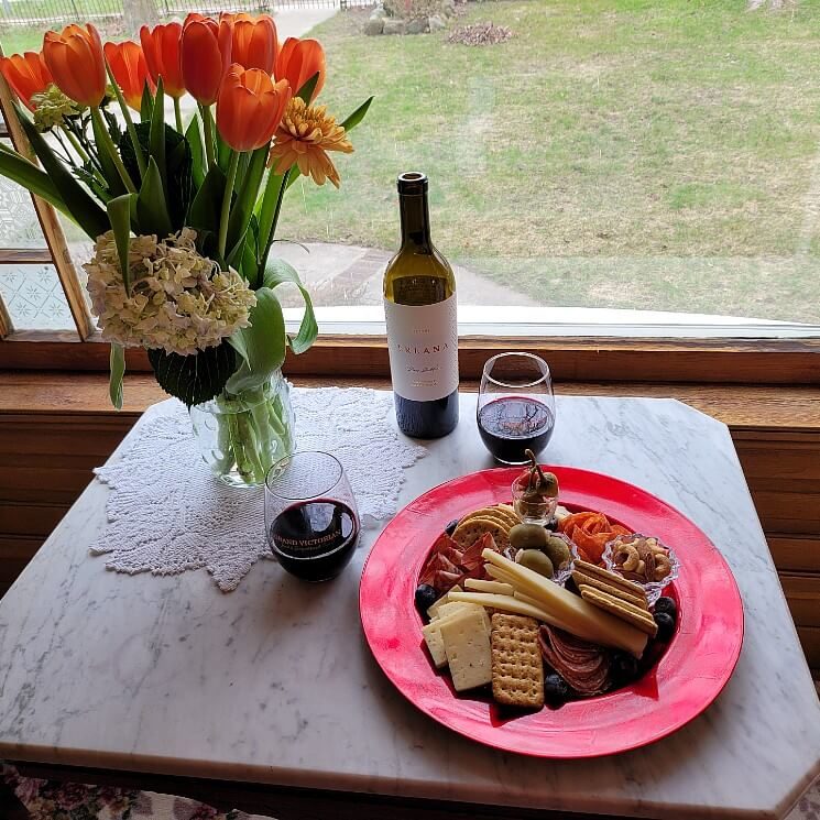 Small white table in front of a window with charcuterie plate, vase of flowers, bottle of wine and two wine glasses