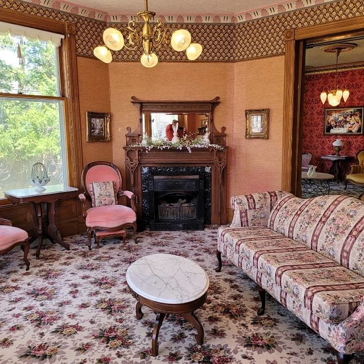 Bright sitting room of a historic home with couch, two chairs, fireplace and doorway into a second room