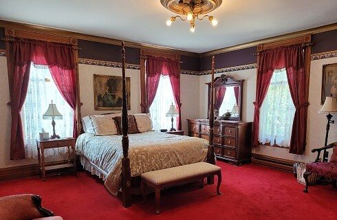 Elegant bedroom in ivory and red with four poster bed, dresser and large curtained windows