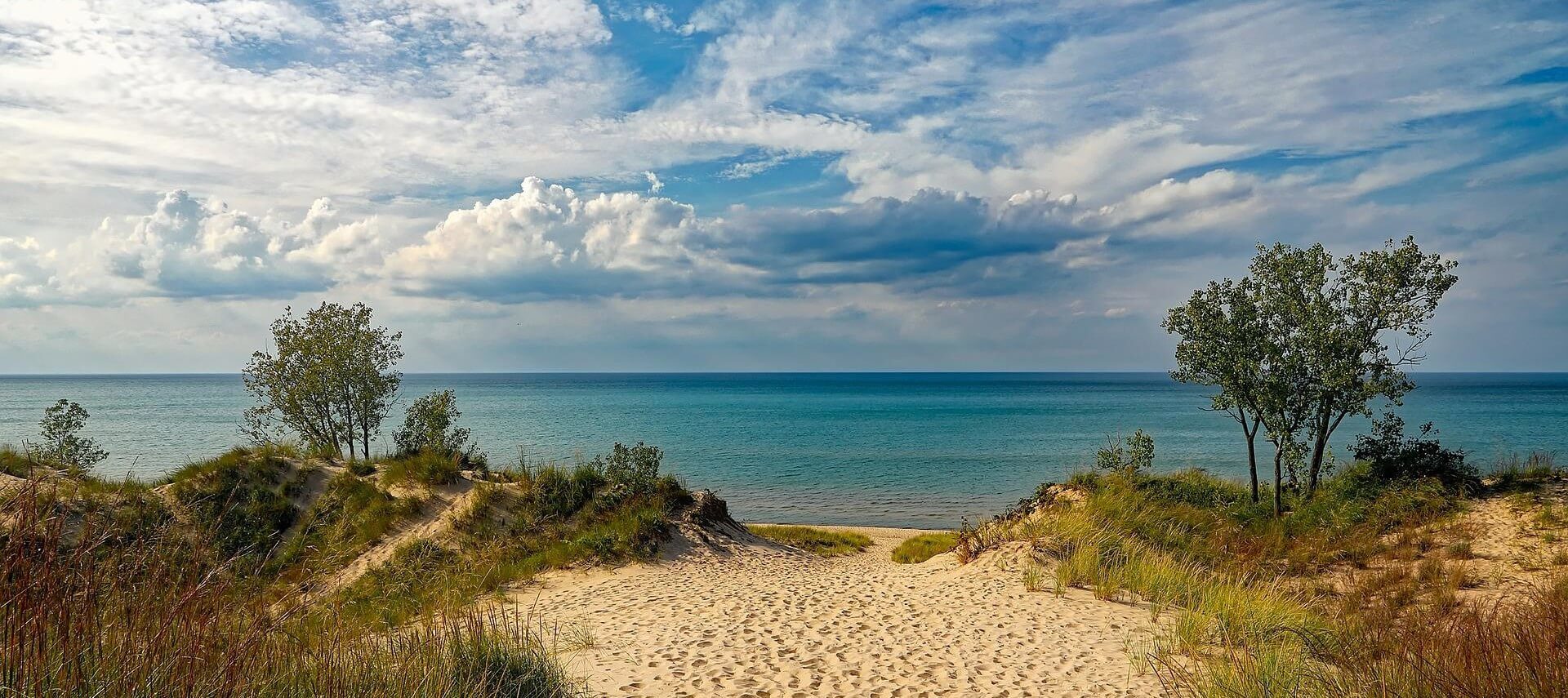 Sandy path leading to an open body of water with clouds and blue sky overhead