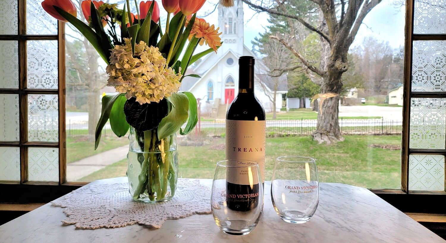 Small white table in front of a window with a bottle of wine, two glasses and a vase of flowers