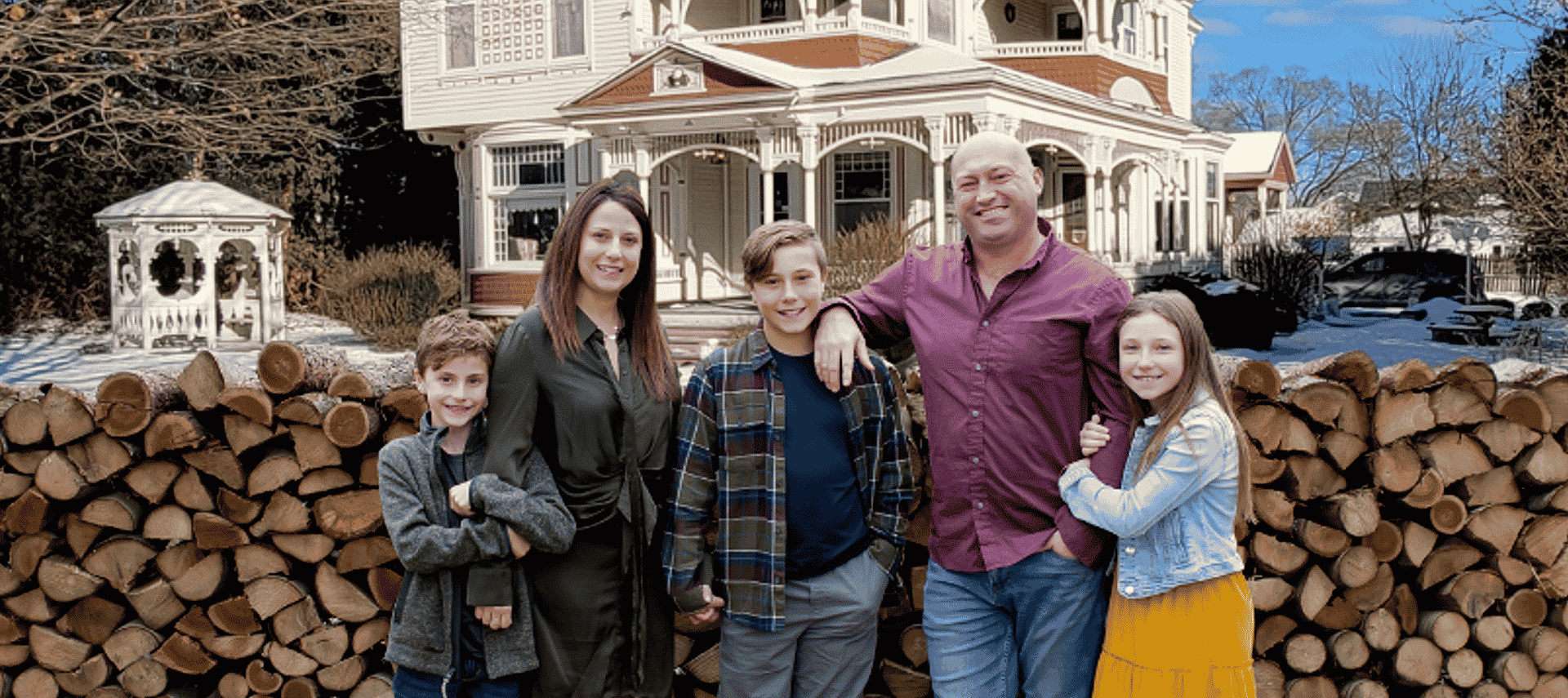 A family with husband, wife and three kids standing in front of a large cord of wood by a large home