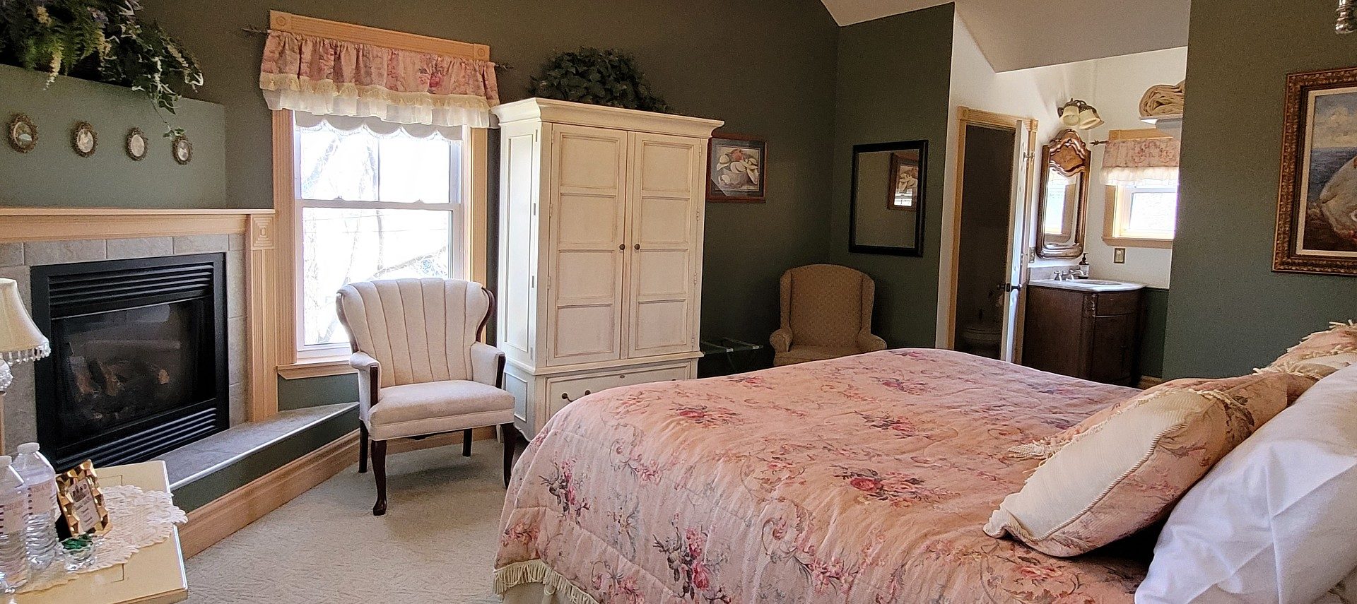 Spacious bedroom with gas fireplace, white armoire, bathroom nook and bright windows
