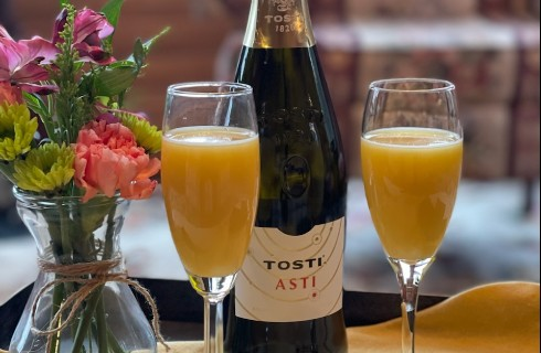 Bottle of champagne next to two glasses of mimosa and a vase of flowers