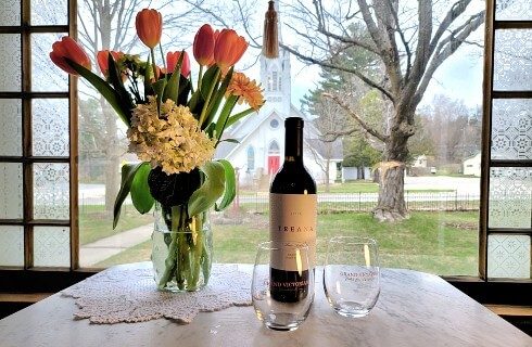 Small white table in front of a window with a bottle of wine, two glasses and a vase of flowers
