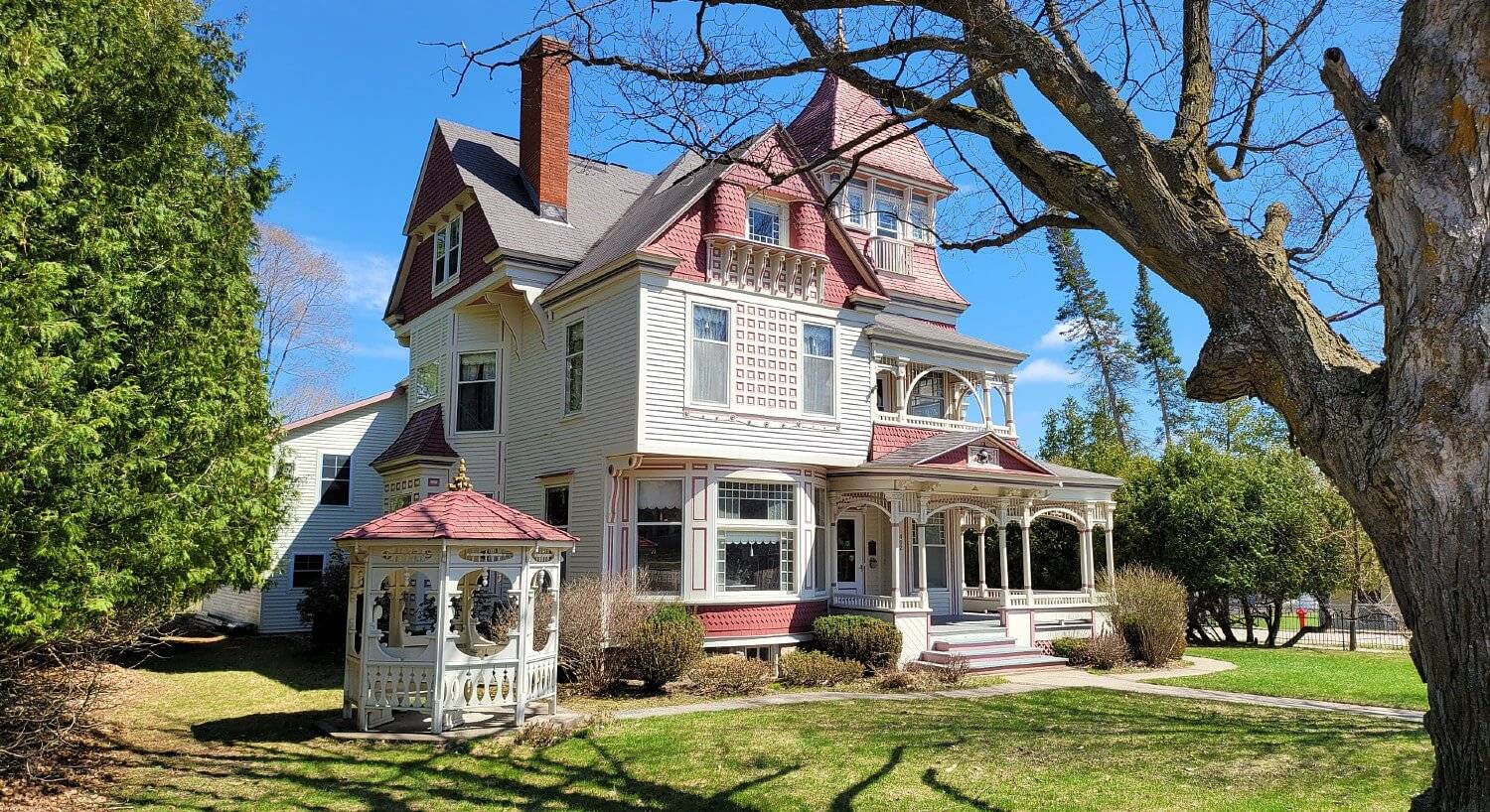 Outdoor view of a large red and white historic home with wrap around porch and surrounded by trees